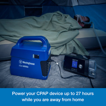 Westinghouse | iGen600s Portable Power Station is shown on the floor of a tent powering a CPAP machine. A man wearing the sleep apnea mask is sleeping on an air mattress in the background. A blue banner along the bottom reads, "Power your CPAP device up to 27 hours while you are away from home".