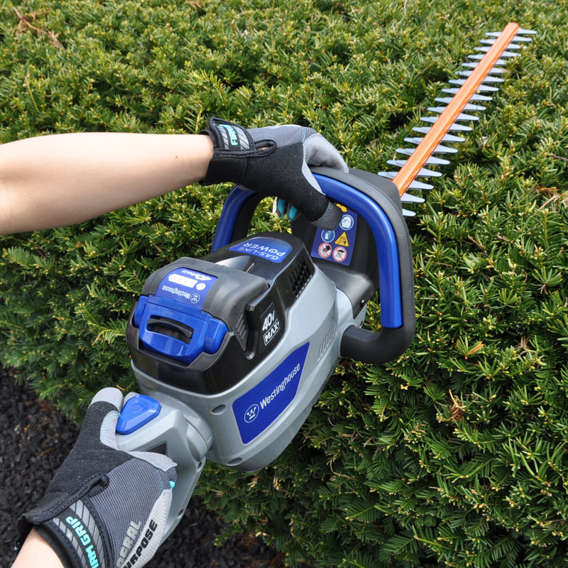 A person holds a hedge trimmer up to a bush. The view is from the rear of the hedge trimmer so the handles are in the foreground while the blades are in the background