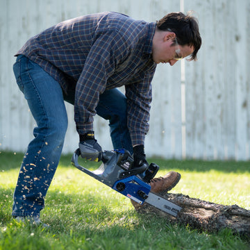 A man holds a tree branch down on the grass with his boot. He is cutting through the branch with a chain saw
