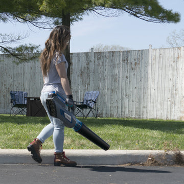A woman walks along the edge of a street using the leaf blower to blow debris off of the street. The background is a yard with a white fence, a tree, and some chairs.